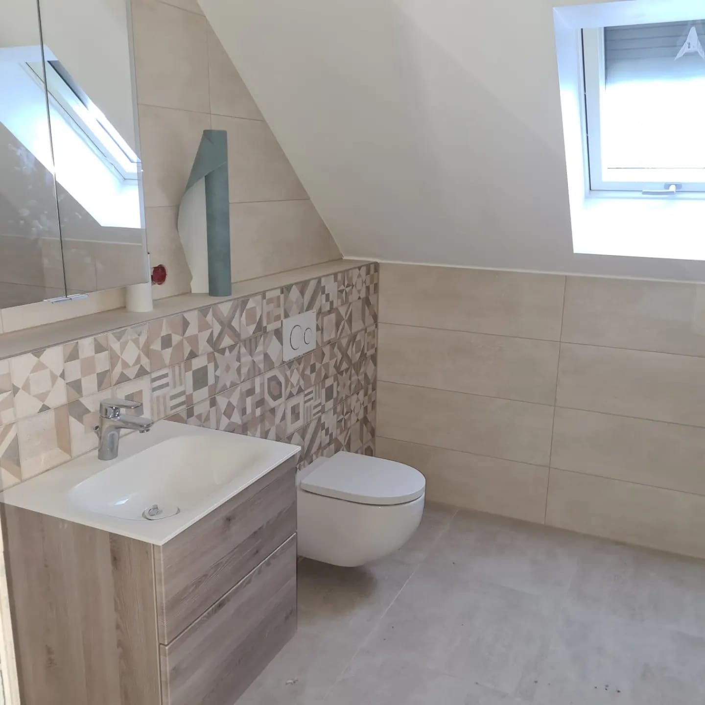 Jasna łazienka na poddaszu z umywalką w szafce z drewnianym dekorem, toaletą i mozaiką na ścianie. Okno dachowe wpuszcza światło.