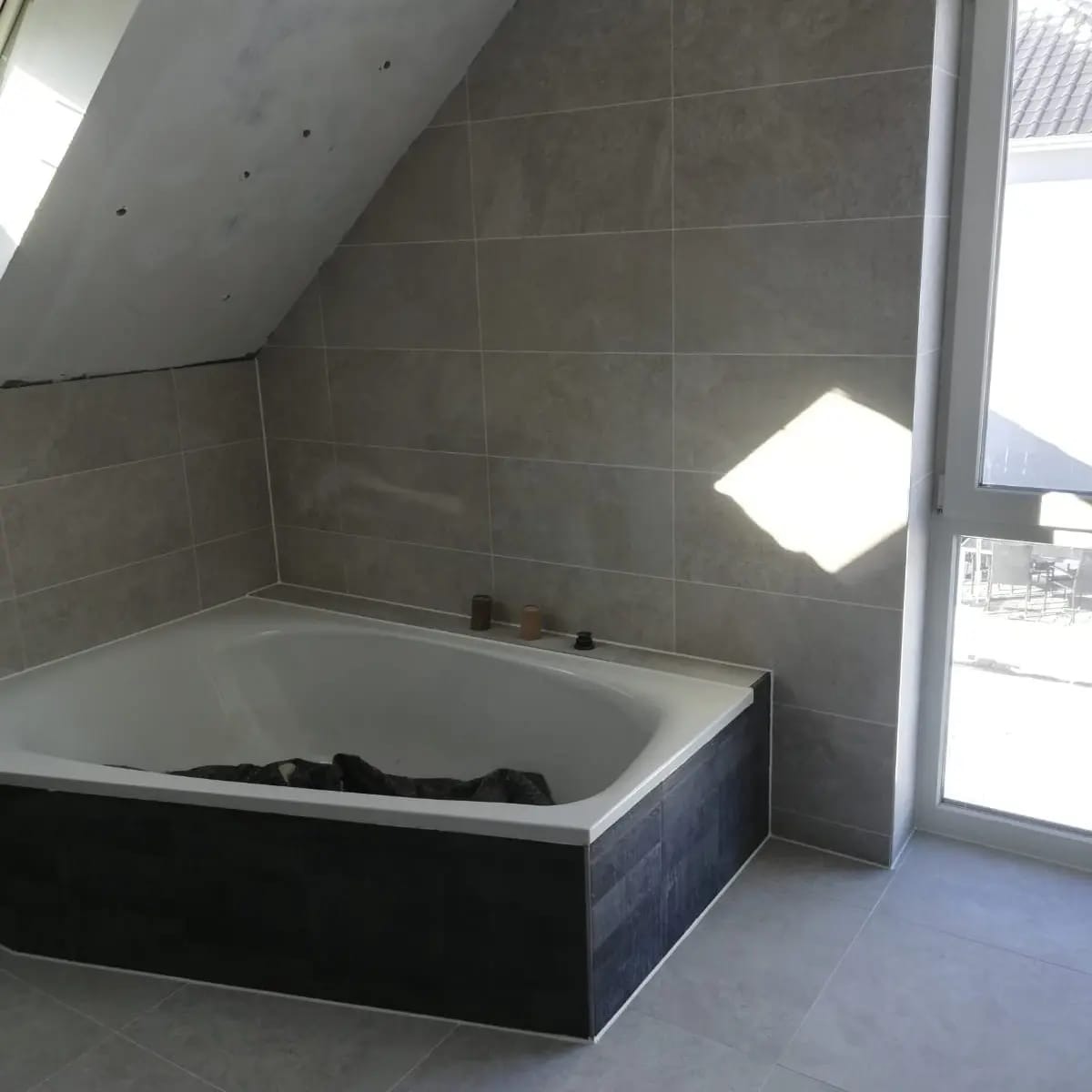 Wykończona łazienka na poddaszu: szare płytki ścienne i podłogowe, biała wanna z czarną obudową, okno z widokiem na zewnątrz. Minimalistyczny design.