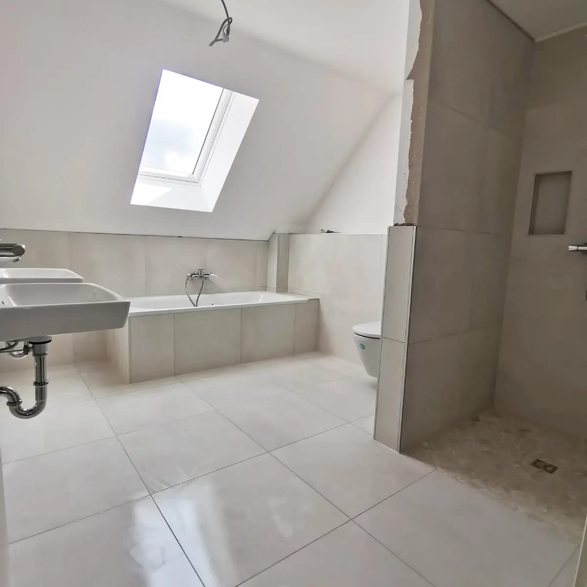 Jasna łazienka na poddaszu z oknem dachowym, wykończona płytkami w odcieniach bieli i szarości. Widoczne umywalki, wanna i kabina prysznicowa.