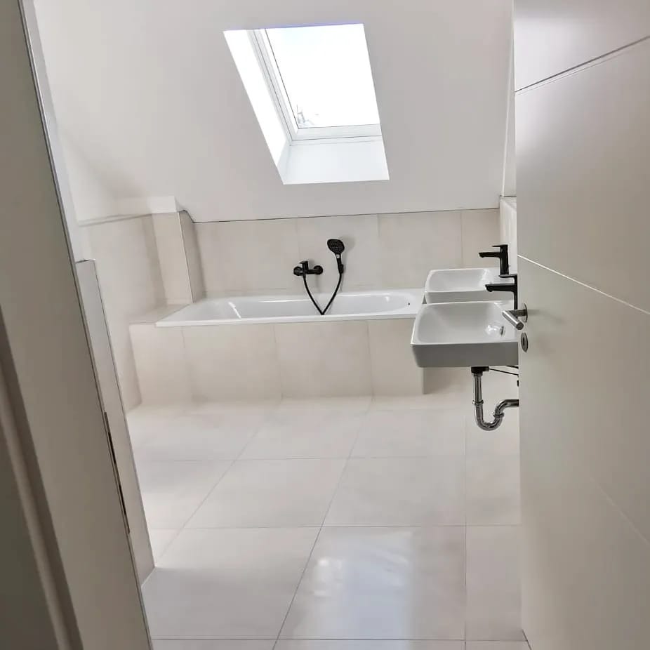 Jasna łazienka na poddaszu z oknem dachowym, wanną obudowaną płytkami i dwiema umywalkami z czarną armaturą. Widok zza uchylonych drzwi.