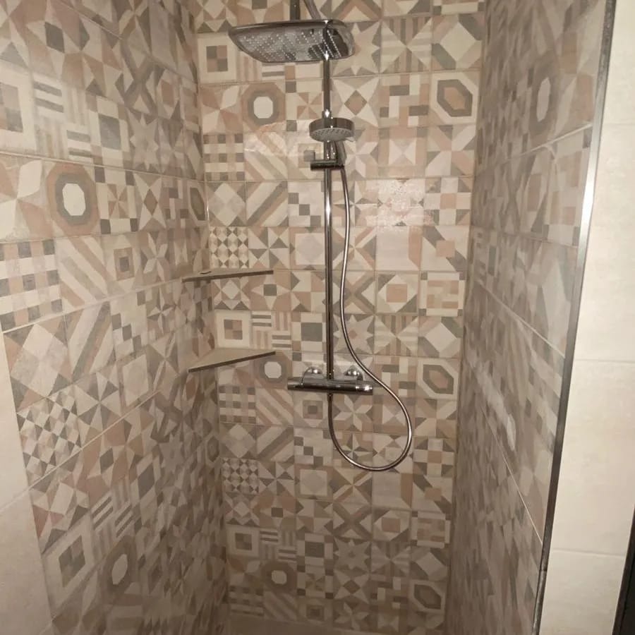 Ukończona kabina prysznicowa z mozaikowymi płytkami w odcieniach beżu i szarości, wyposażona w deszczownicę i półki na kosmetyki, wykończona listwami.