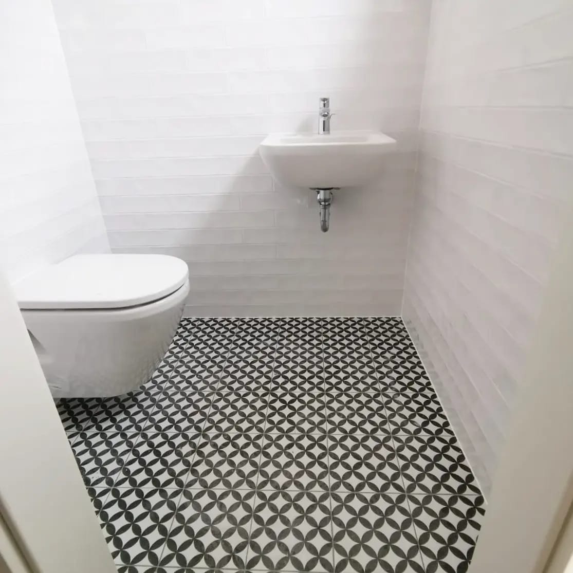 Biała łazienka z umywalką i toaletą. Na podłodze wzorzyste, czarno-białe płytki. Ściany wyłożone białymi, podłużnymi płytkami.