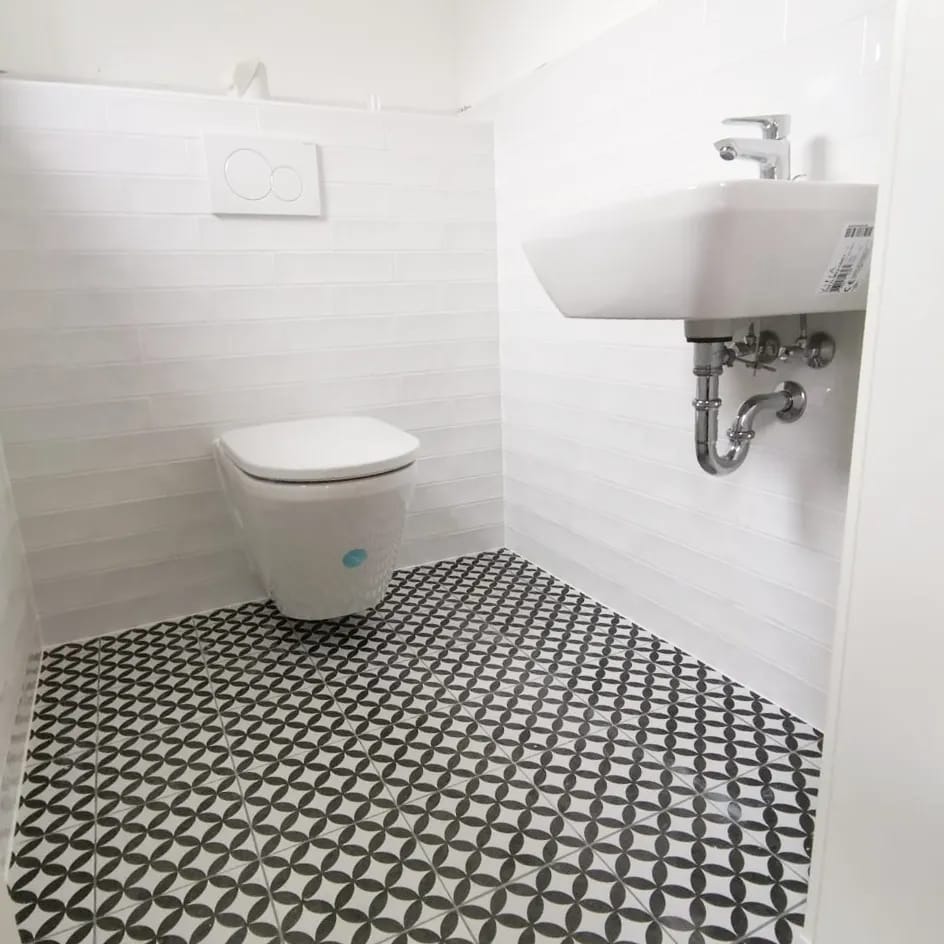 Biała łazienka z umywalką, podwieszaną toaletą i czarno-białą podłogą w geometryczny wzór. Ściany wyłożone białymi, poziomymi płytkami imitującymi cegłę.