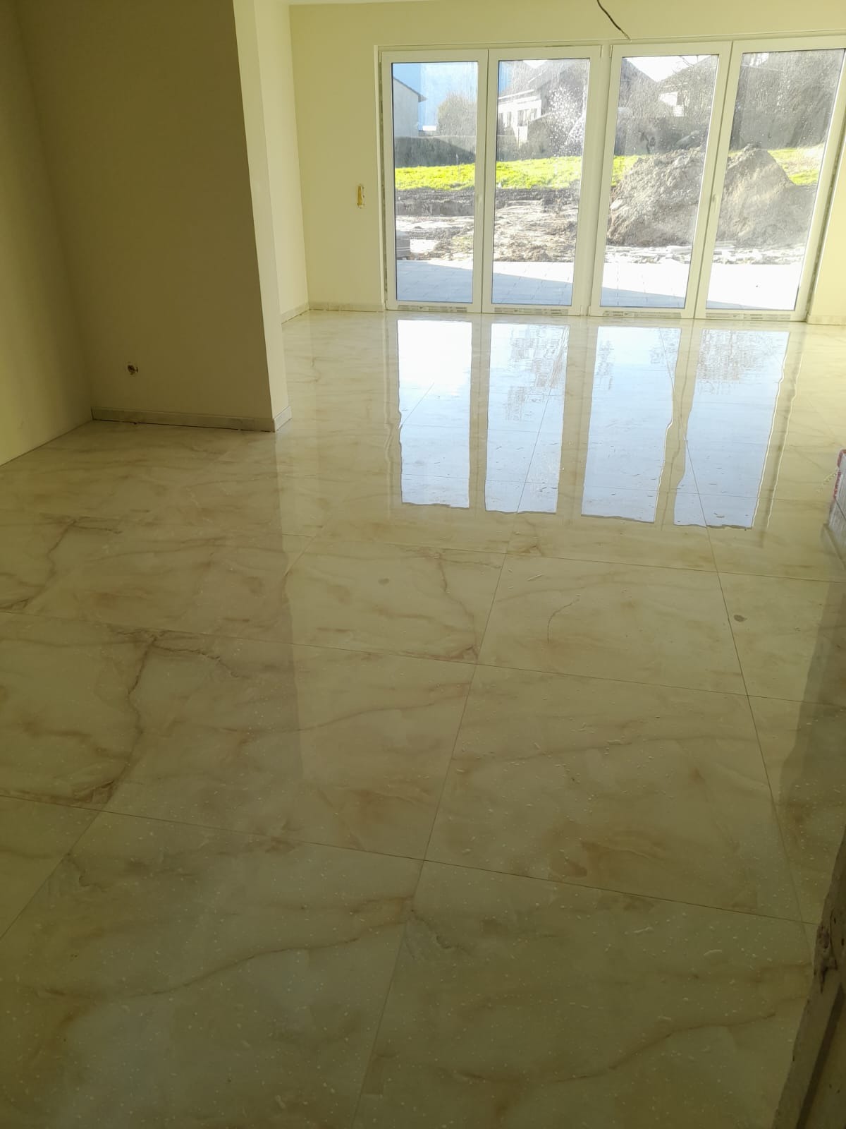 Jasne, marmurkowe płytki podłogowe w nowym pomieszczeniu z dużymi oknami, odbijającymi światło. Widok na zewnątrz, gdzie widać ziemię i zieleń.