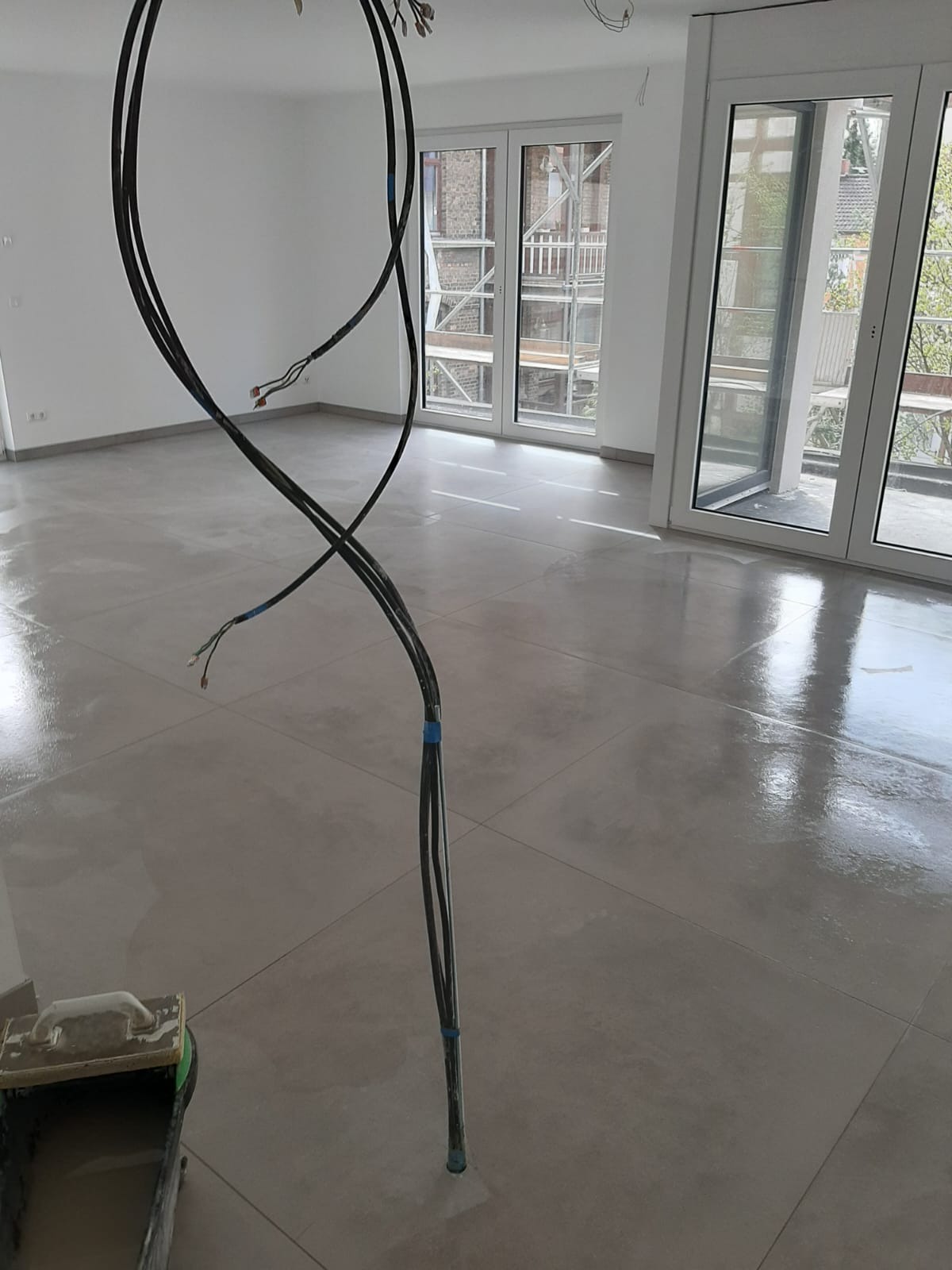 Błyszcząca, jasnoszara podłoga z płytek w nowym, pustym pomieszczeniu. Widoczne wiszące kable i narzędzia glazurnicze. Duże okna balkonowe w tle.