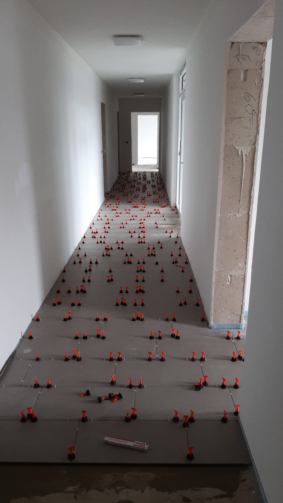 Korytarz w trakcie układania płytek podłogowych. Widoczne szare płyty wyrównane z użyciem pomarańczowych klipsów i klinów. Ściany białe, surowe. W tle widoczne kolejne pomieszczenia.