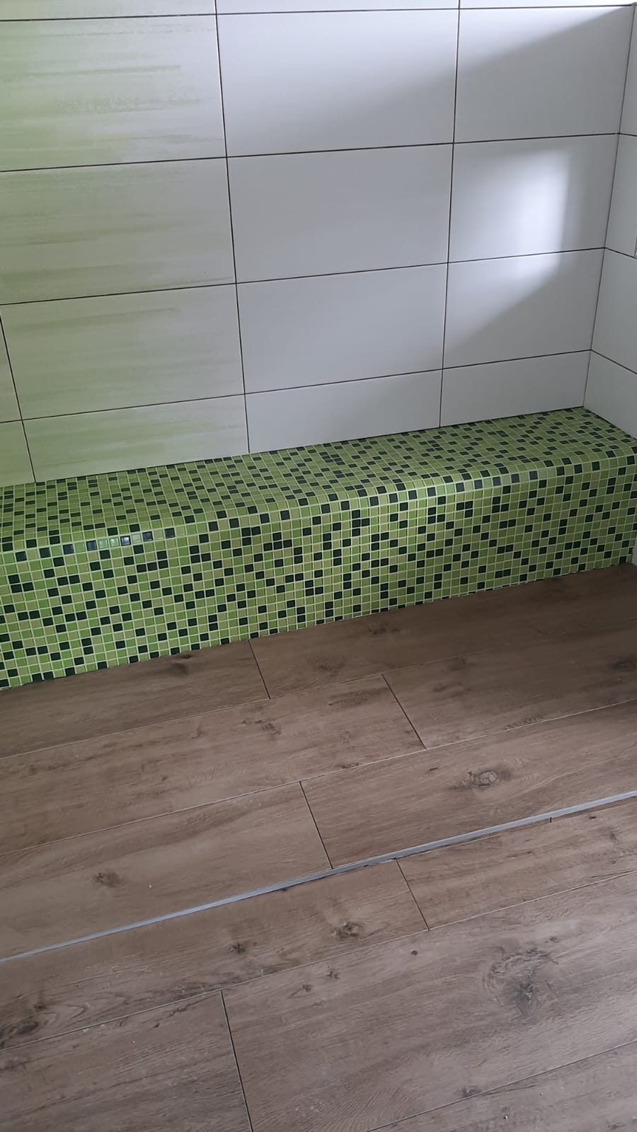 Wykończona łazienka z zieloną mozaiką na obudowie wanny/brodzika, jasne płytki na ścianach i panele podłogowe imitujące drewno. Widok z góry pod kątem.