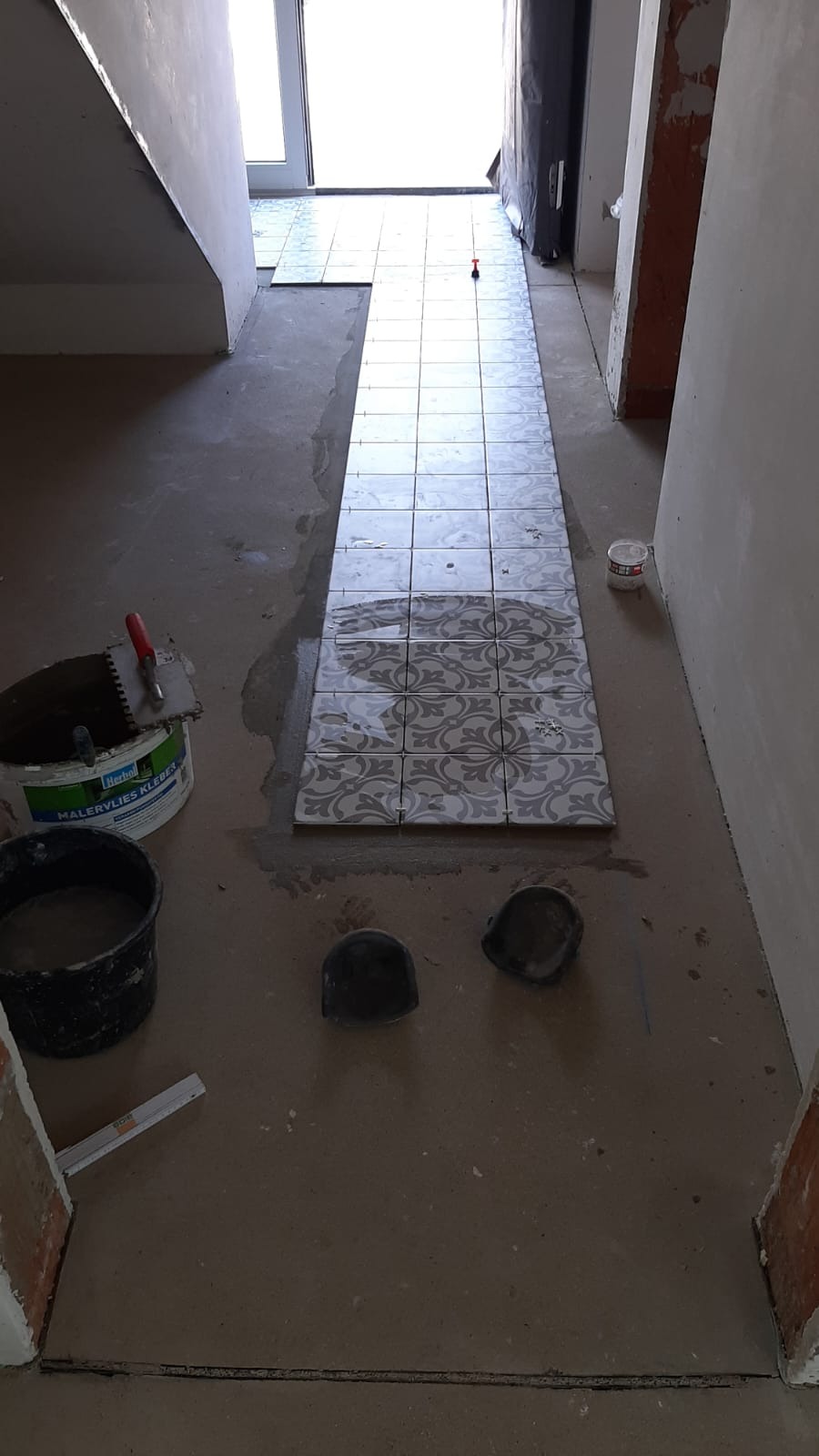 Układanie wzorzystych płytek podłogowych w korytarzu, widoczne narzędzia glazurnicze i wiadro z klejem. Częściowo wyłożona posadzka, surowy stan wykończenia ścian.