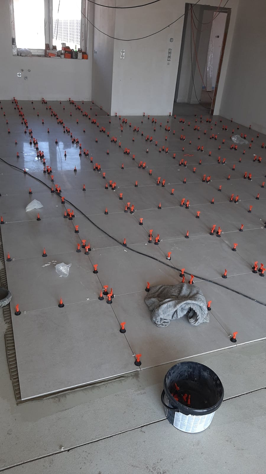 Układanie płytek podłogowych z użyciem systemu poziomowania. Widoczny kubeł z klipsami, szmatka i równo ułożone płytki w szarym odcieniu. W tle surowe ściany w trakcie remontu.