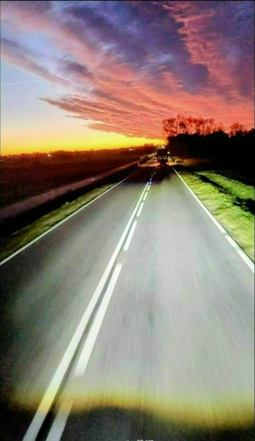 Asfaltowa droga nocą, oświetlona światłami samochodu w oddali, z dramatycznym, kolorowym zachodem słońca nad horyzontem. Długie cienie i intensywne barwy.