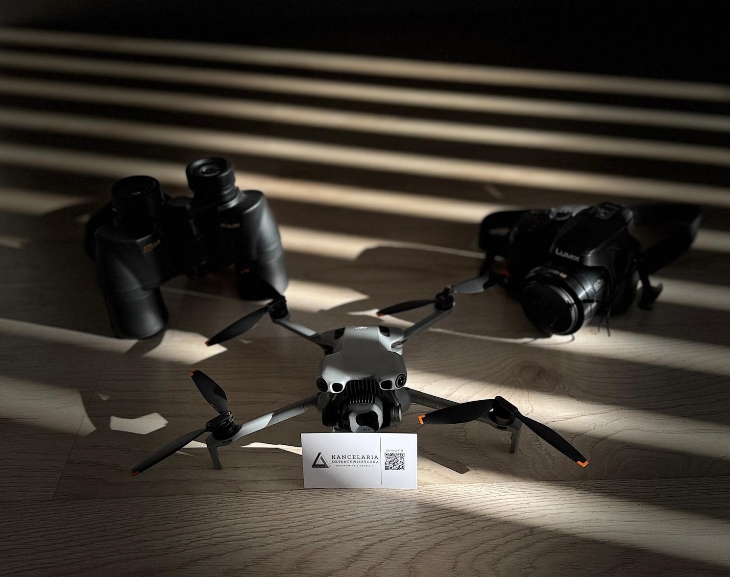 Dron, lornetka i aparat fotograficzny w cieniu na drewnianej podłodze. Wizytówka kancelarii detektywistycznej z kodem QR na pierwszym planie.