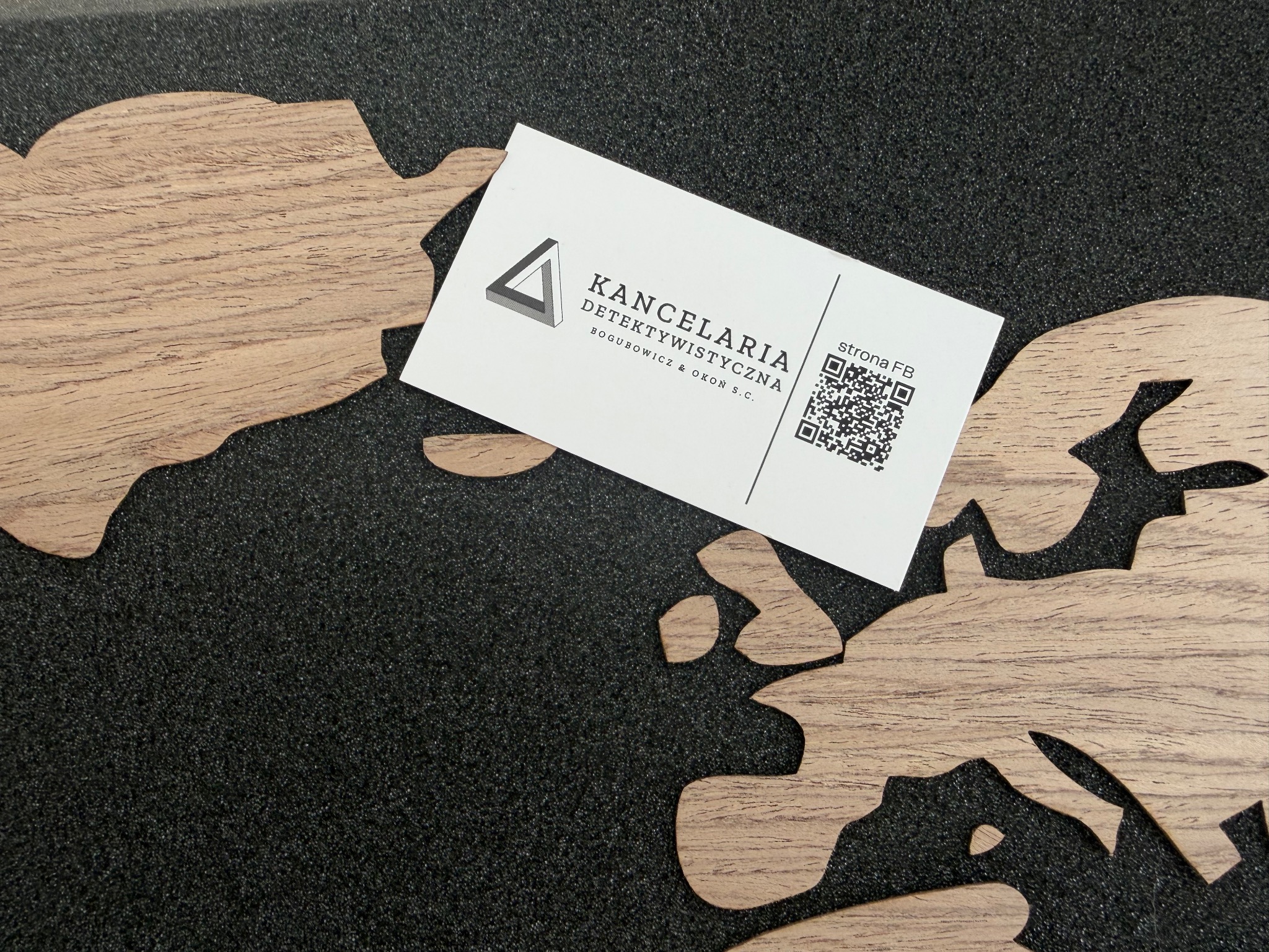 Wizytówka kancelarii detektywistycznej z QR kodem na tle czarnego materiału i drewnianych elementów dekoracyjnych. Minimalistyczny design, stonowana kolorystyka.