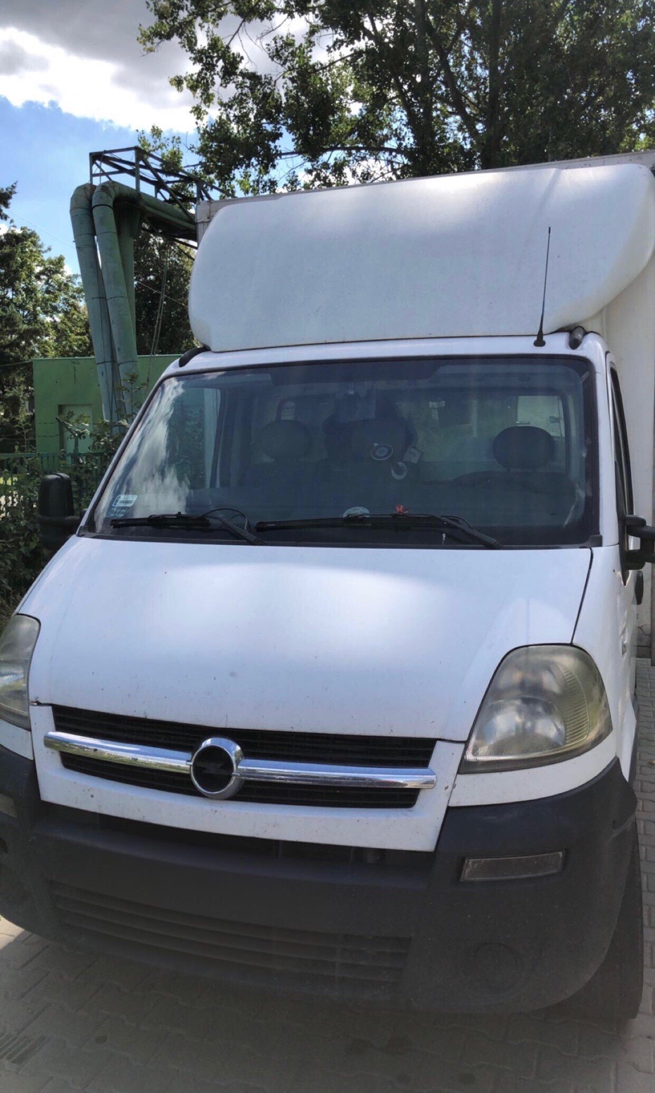 Biały samochód dostawczy z wysoką zabudową, zaparkowany na zewnątrz, z elementami przemysłowymi w tle i drzewami. Widok z przodu, lekko z dołu.