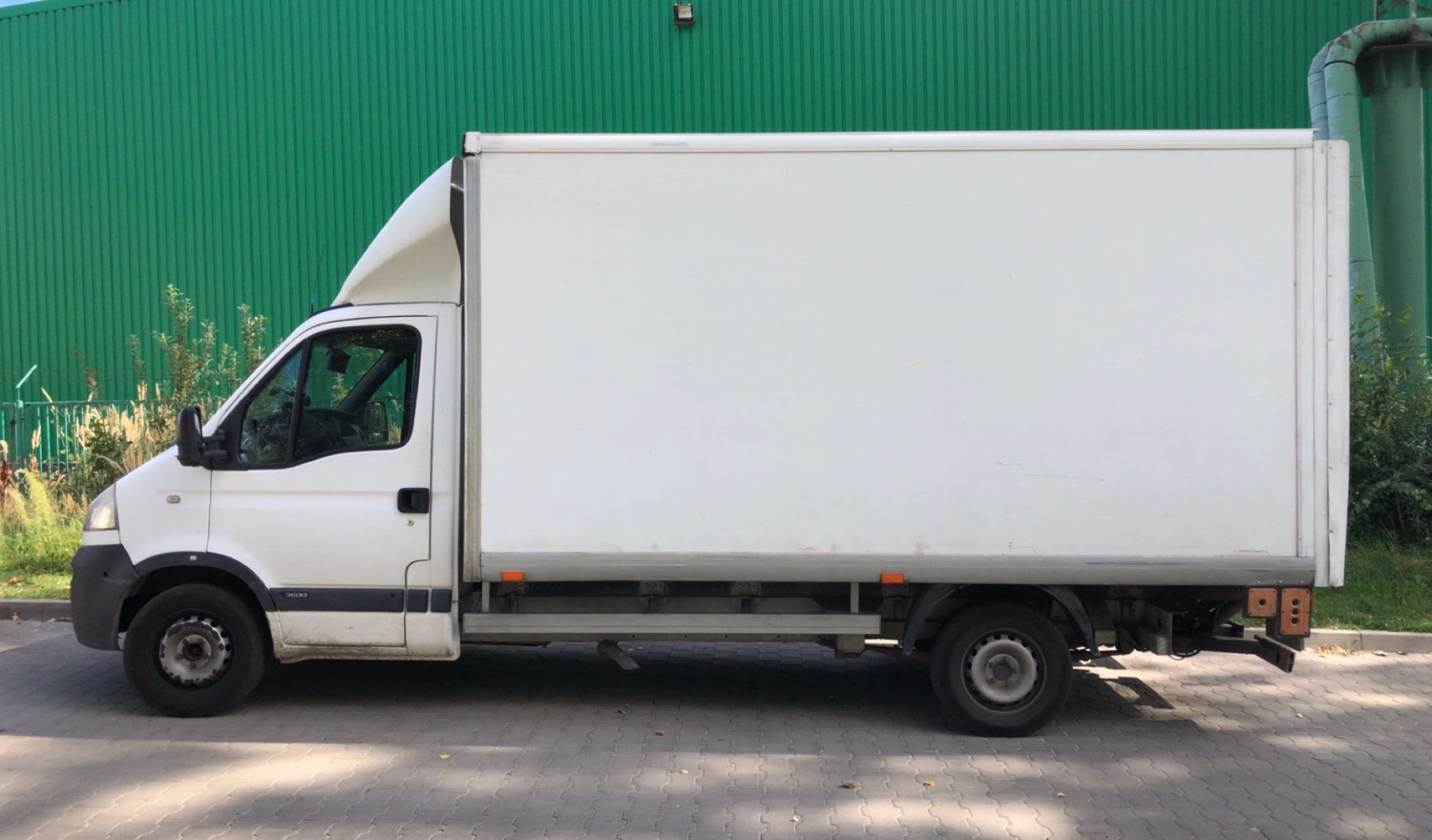 Biała furgonetka z pustą przestrzenią ładunkową na tle zielonej ściany. Widok z boku, światło dzienne.