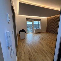 Harmonia Katow - Jasne parkiety, drewniana ściana z lamelami i podświetlany sufit w przestronnym pokoju z balkonem. Widoczny domofon i włącznik na ścianie.