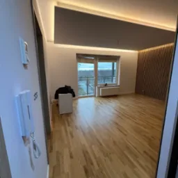 Jasne parkiety, drewniana ściana z lamelami i podświetlany sufit w przestronnym pokoju z balkonem. Widoczny domofon i włącznik na ścianie.