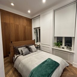 Harmonia Katow - Sypialnia po remoncie: parkiet w jodełkę, drewniana zabudowa ściany z szafami, okna z roletami i roślinami, łóżko z zielonym pledem. Przytulne wykończenie wnętrza.