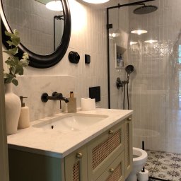 Harmonia Katow - Stylowa łazienka z kabiną prysznicową typu walk-in, umywalką z szafką w odcieniach zieleni i okrągłym lustrem. Białe płytki i wzorzysta podłoga dodają elegancji.