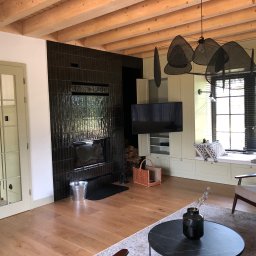 Harmonia Katow - Nowoczesny salon z drewnianą podłogą, czarnym kominkiem z błyszczących płytek i oknem z żaluzjami. Widoczne drzwi z przeszkleniami i drewniany sufit z belkami.