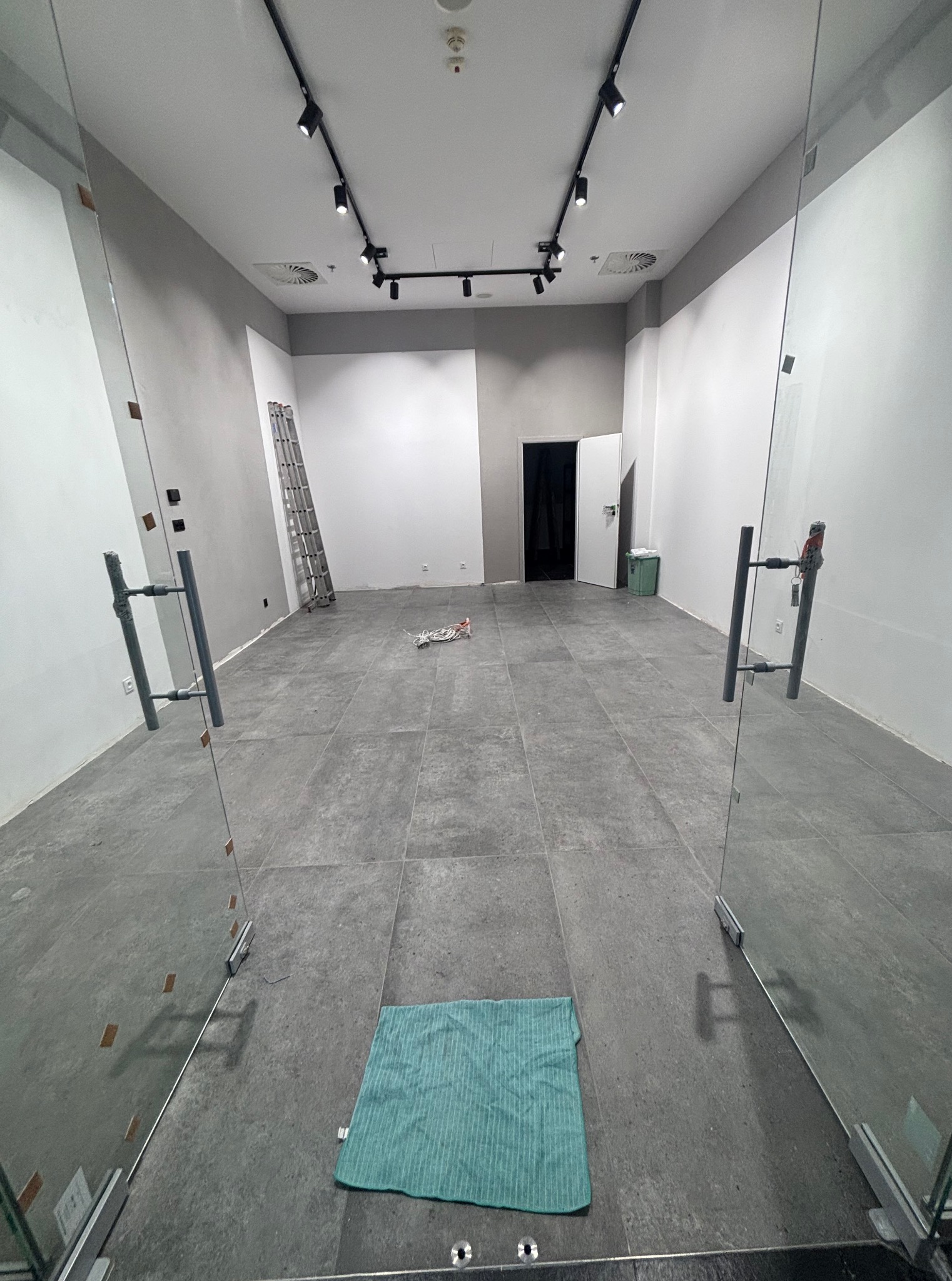 Wnętrze biura po remoncie: szare płytki podłogowe, białe ściany z szarym pasem, szklane drzwi wejściowe, drabina oparta o ścianę. Widoczne ślady po pracach wykończeniowych.