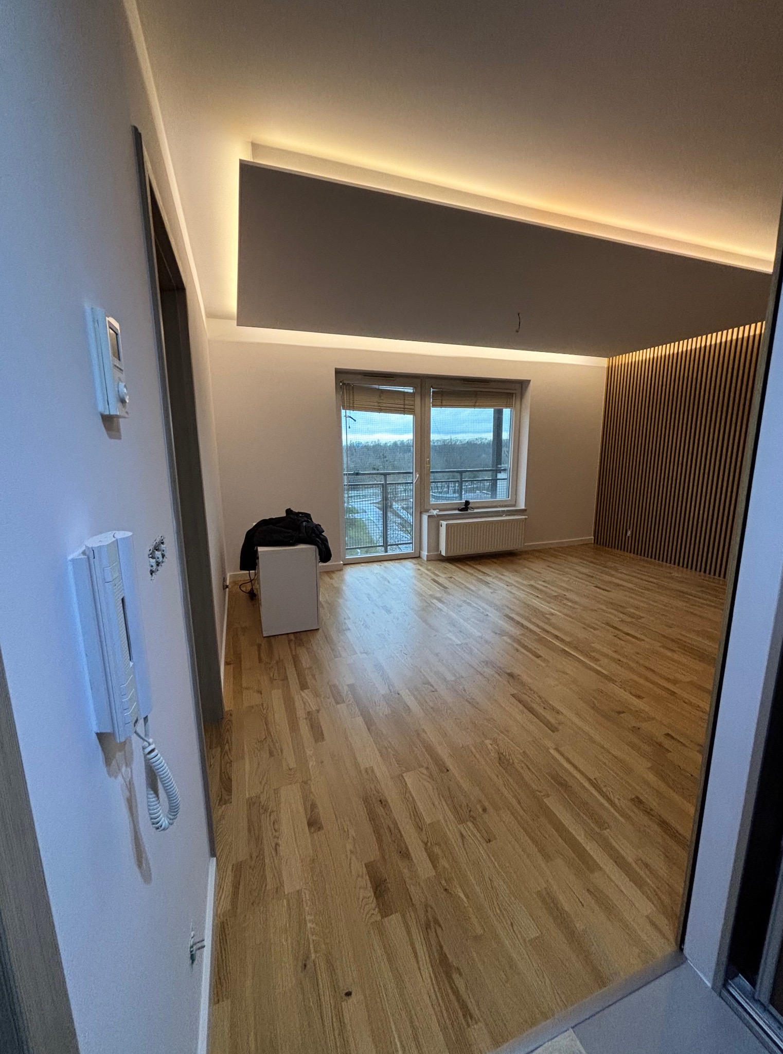 Jasne parkiety, drewniana ściana z lamelami i podświetlany sufit w przestronnym pokoju z balkonem. Widoczny domofon i włącznik na ścianie.