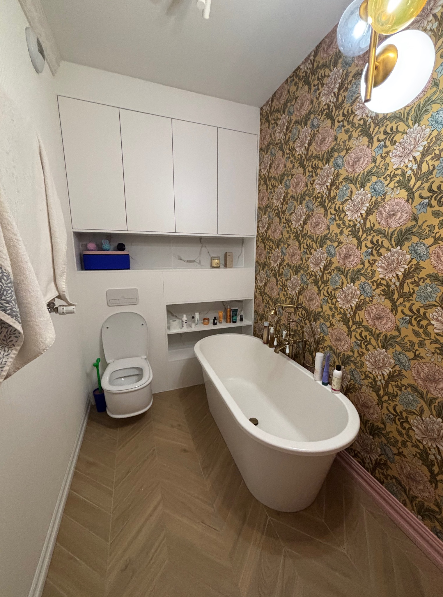 Elegancka łazienka po remoncie: wolnostojąca wanna, toaleta, szafki w zabudowie i wzorzysta tapeta w kwiaty. Podłoga wyłożona parkietem w jodełkę.