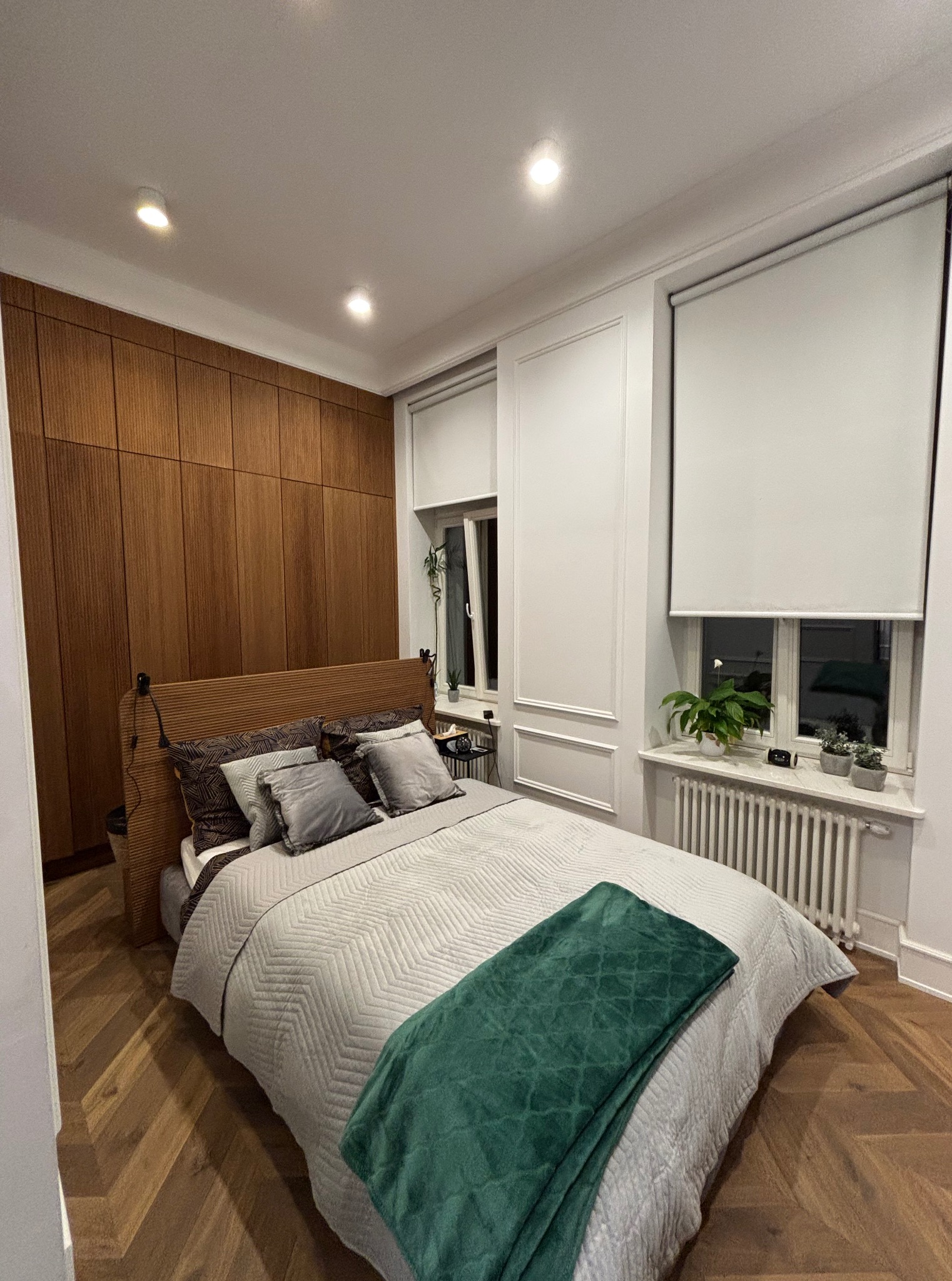 Sypialnia po remoncie: parkiet w jodełkę, drewniana zabudowa ściany z szafami, okna z roletami i roślinami, łóżko z zielonym pledem. Przytulne wykończenie wnętrza.