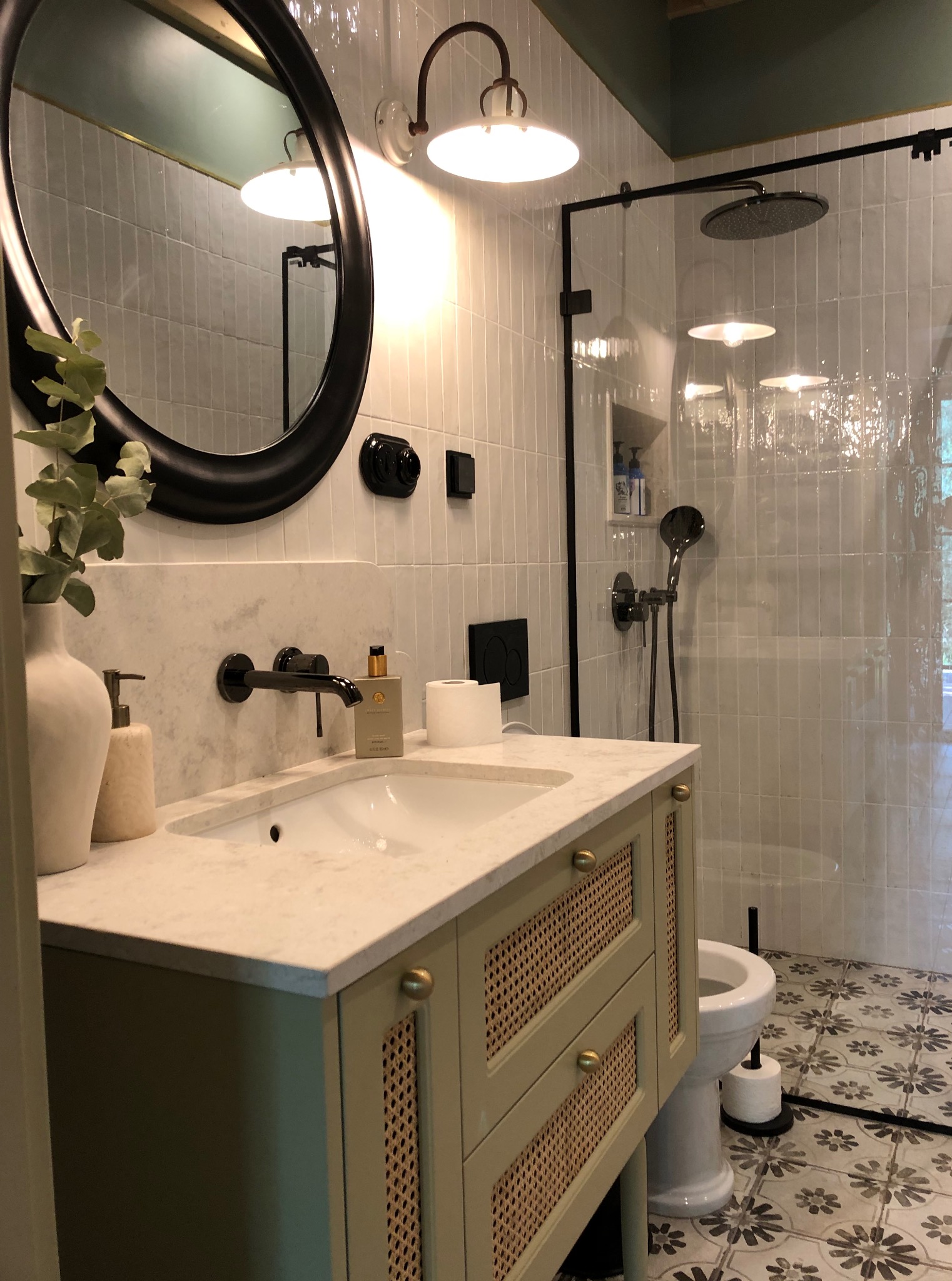 Stylowa łazienka z kabiną prysznicową typu walk-in, umywalką z szafką w odcieniach zieleni i okrągłym lustrem. Białe płytki i wzorzysta podłoga dodają elegancji.