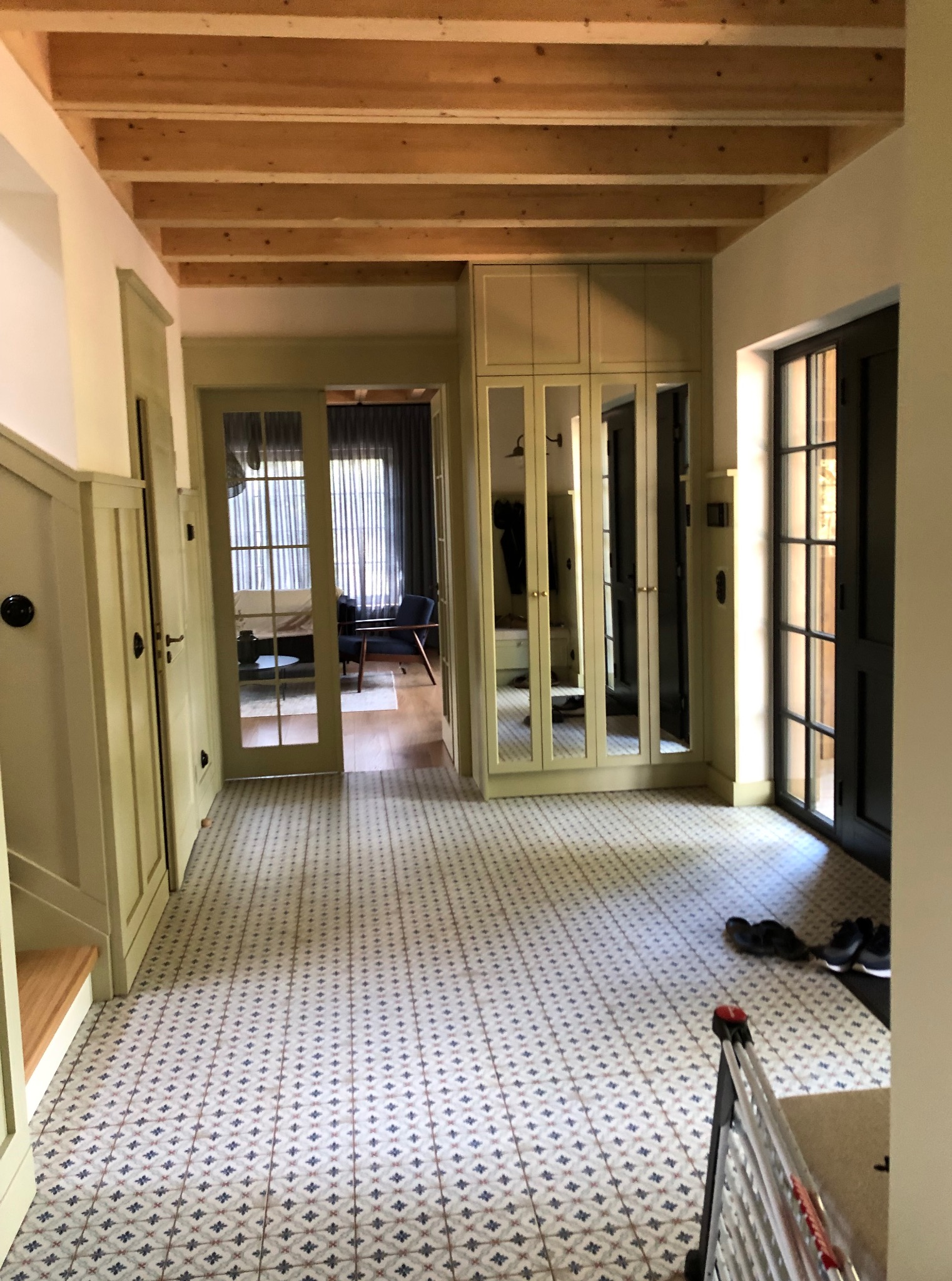 Jasny korytarz z wzorzystą podłogą i drewnianym sufitem. Widoczne drzwi, szafa z lustrami i fragment drabiny malarskiej. Ukończone wykończenie wnętrza.