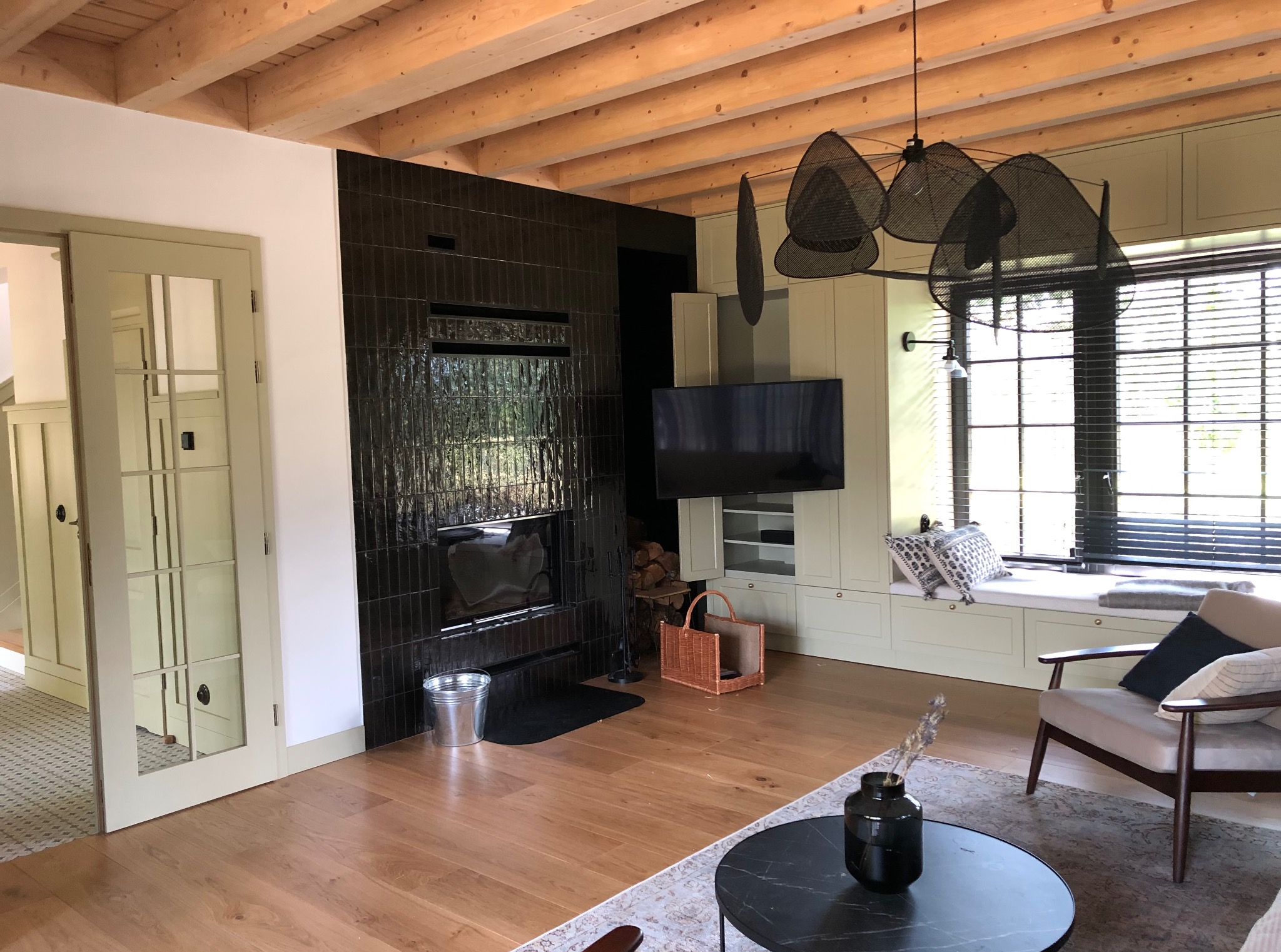 Nowoczesny salon z drewnianą podłogą, czarnym kominkiem z błyszczących płytek i oknem z żaluzjami. Widoczne drzwi z przeszkleniami i drewniany sufit z belkami.