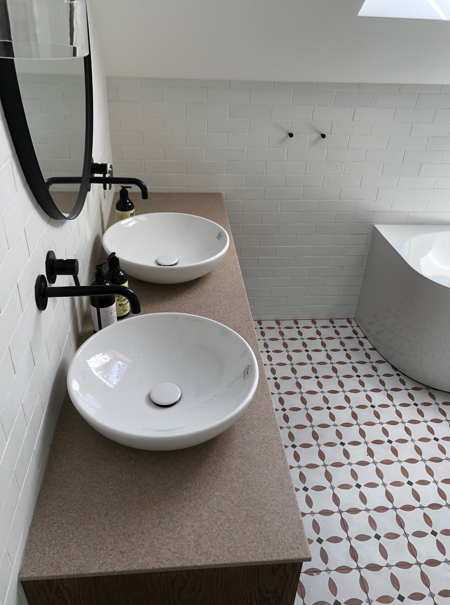 Biała łazienka z dwoma umywalkami nablatowymi, czarną armaturą i mozaiką na podłodze. Widoczna wanna i okno dachowe. Minimalistyczny design.