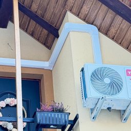 Klimmar - Biały klimatyzator TCL zamontowany na elewacji domu z drewnianą podbitką, połączony białymi rurami z wnętrzem. Widoczny wieniec na drzwiach i kwiaty w doniczce.
