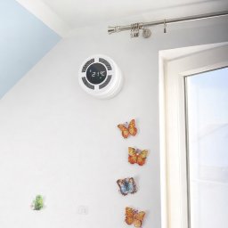 Klimmar - Ścienna jednostka rekuperacji bez centrali, z wyświetlaczem temperatury, zamontowana na białej ścianie w pokoju na poddaszu, obok okna i dekoracji w kształcie motyli.