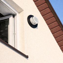 Klimmar - Biała kratka wentylacyjna z czarnym kołnierzem na elewacji domu, obok okna i pod brązowym dachem. Widok z dołu na błękitne niebo.