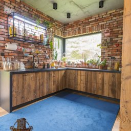 Holy-Wood - Rustykalna kuchnia z drewnianymi szafkami, ceglaną ścianą i niebieskim dywanem. Nad blatem półka z ziołami i oknem. Widoczne oświetlenie punktowe.