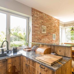 Holy-Wood - Rustykalna kuchnia z drewnianymi frontami, granitowym blatem i ceglaną ścianą. Widok na okno z roślinami i akcesoria kuchenne na blacie.