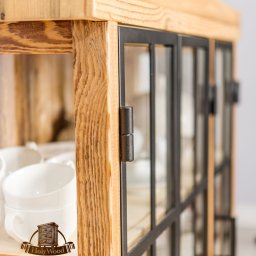 Holy-Wood - Drewniana witryna z metalowymi ramkami, widoczne detale usłojenia i zawiasy. Wewnątrz białe filiżanki i logo producenta. Mebel w stylu rustykalnym.