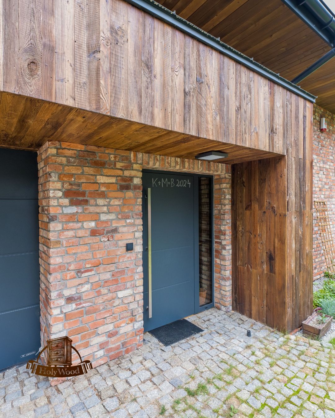 Nowoczesne drzwi wejściowe w domu z cegły i drewna. Na drzwiach napis K+M+B 2024. Elewacja z drewnianych desek. Brukowana ścieżka.
