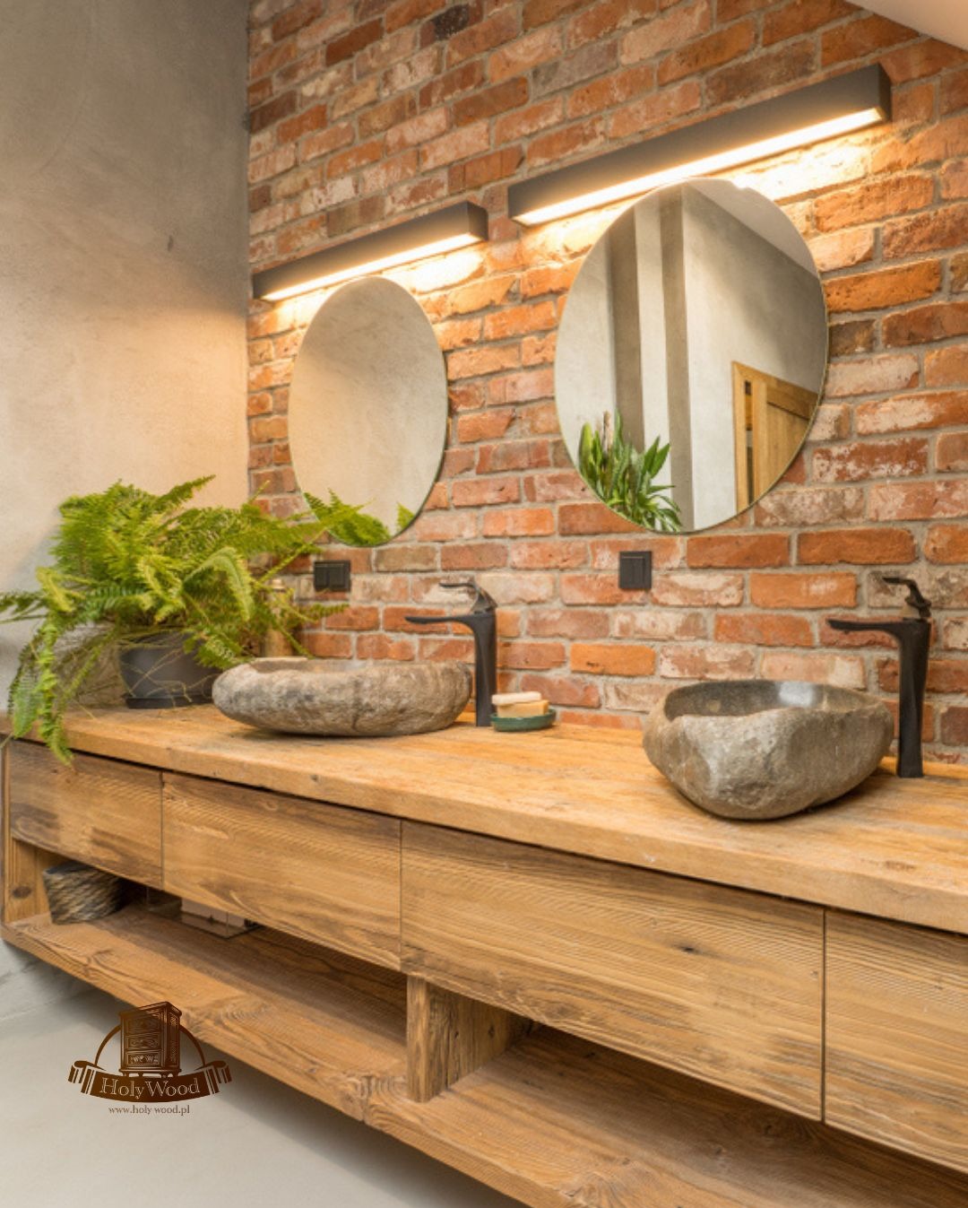 Rustykalna łazienka z drewnianą szafką pod umywalki z kamienia, okrągłe lustra i ceglana ściana. Roślina doniczkowa dodaje naturalnego akcentu.