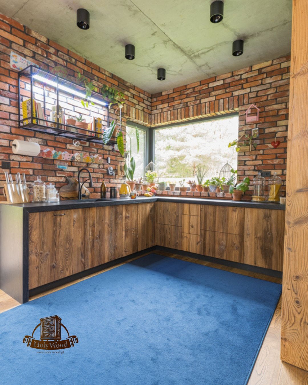 Rustykalna kuchnia z drewnianymi szafkami, ceglaną ścianą i niebieskim dywanem. Nad blatem półka z ziołami i oknem. Widoczne oświetlenie punktowe.