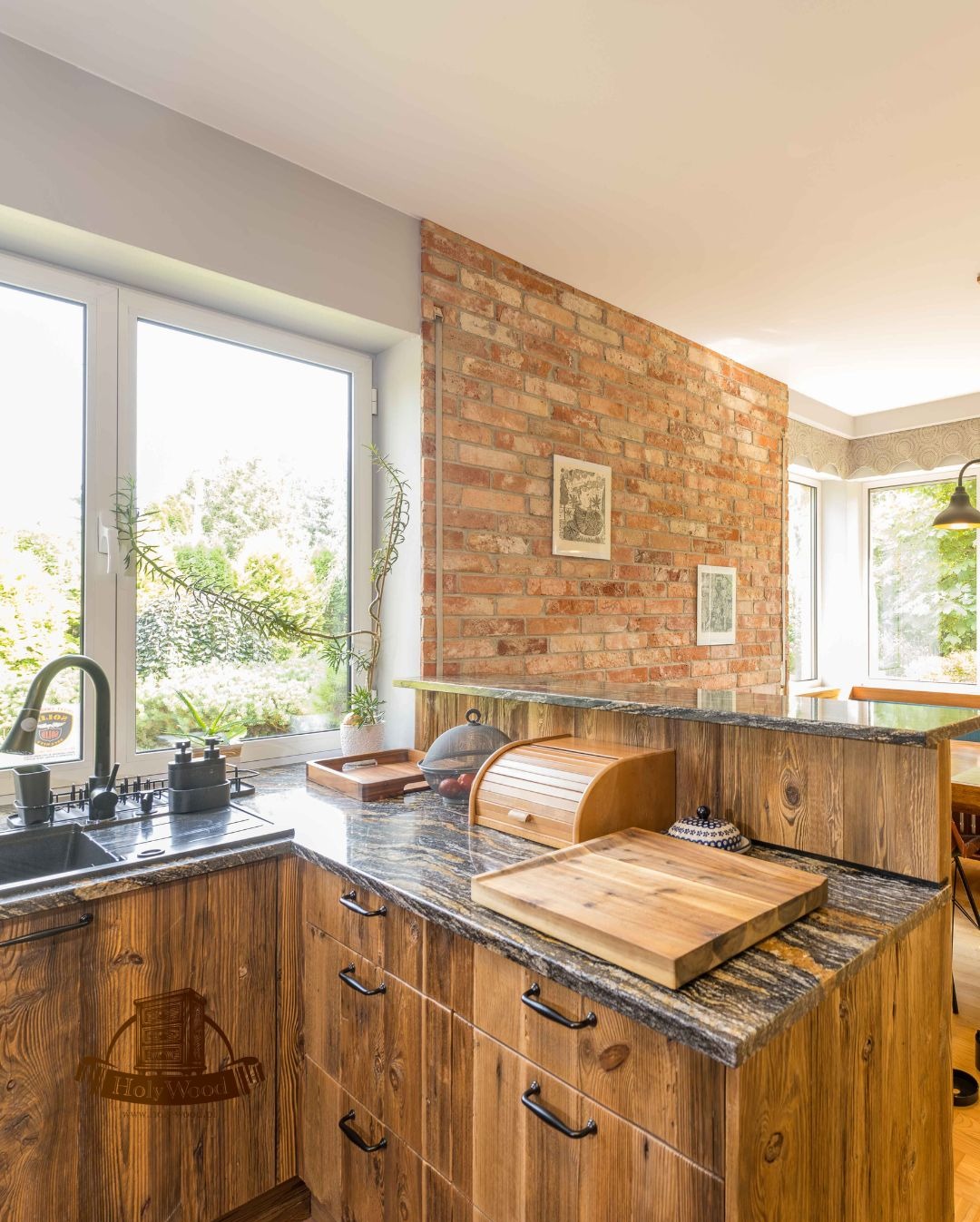 Rustykalna kuchnia z drewnianymi frontami, granitowym blatem i ceglaną ścianą. Widok na okno z roślinami i akcesoria kuchenne na blacie.