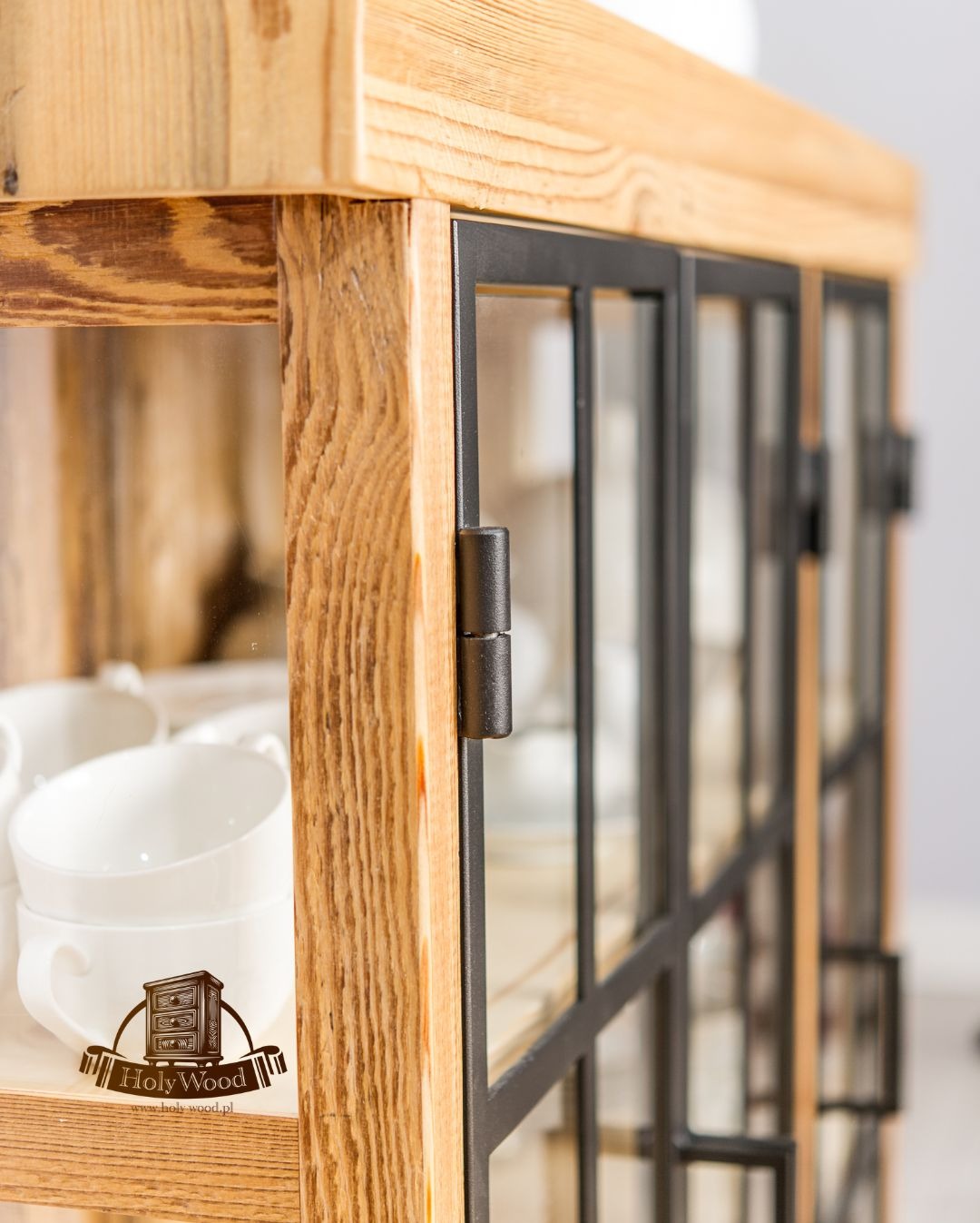 Drewniana witryna z metalowymi ramkami, widoczne detale usłojenia i zawiasy. Wewnątrz białe filiżanki i logo producenta. Mebel w stylu rustykalnym.