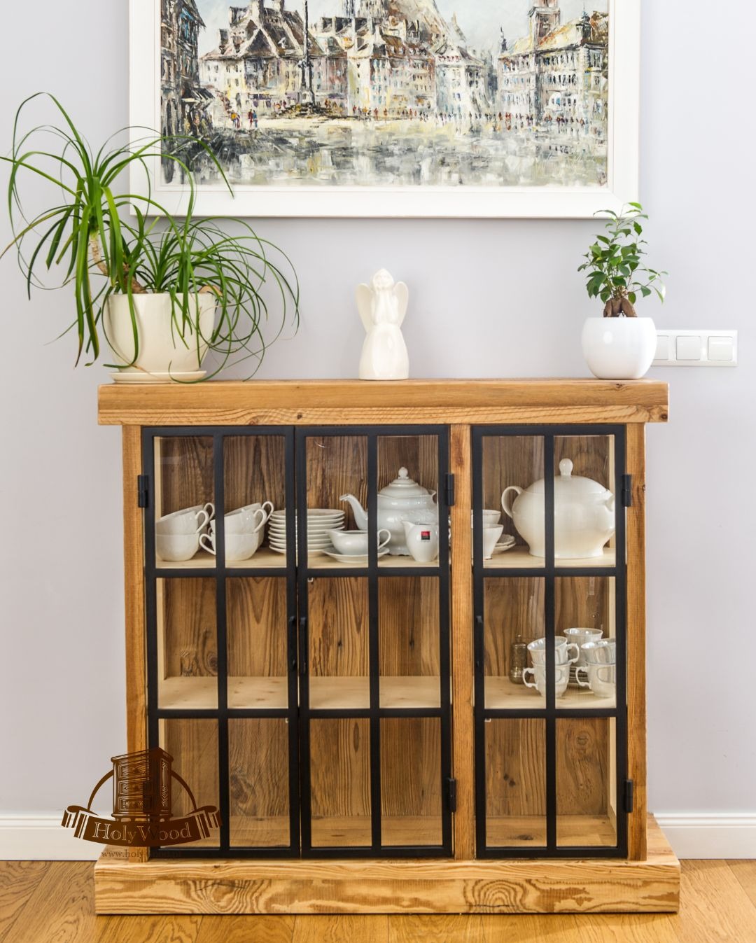 Drewniana witryna z czarnymi szprosami, wypełniona białą porcelaną. Nad nią obraz z miejskim pejzażem, rośliny w doniczkach i figurka anioła.