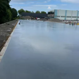 Świeżo wylana, gładka posadzka przemysłowa na zewnątrz budynku, z widocznym sprzętem budowlanym i przygotowanym podłożem. W tle budynek przemysłowy i drzewa.