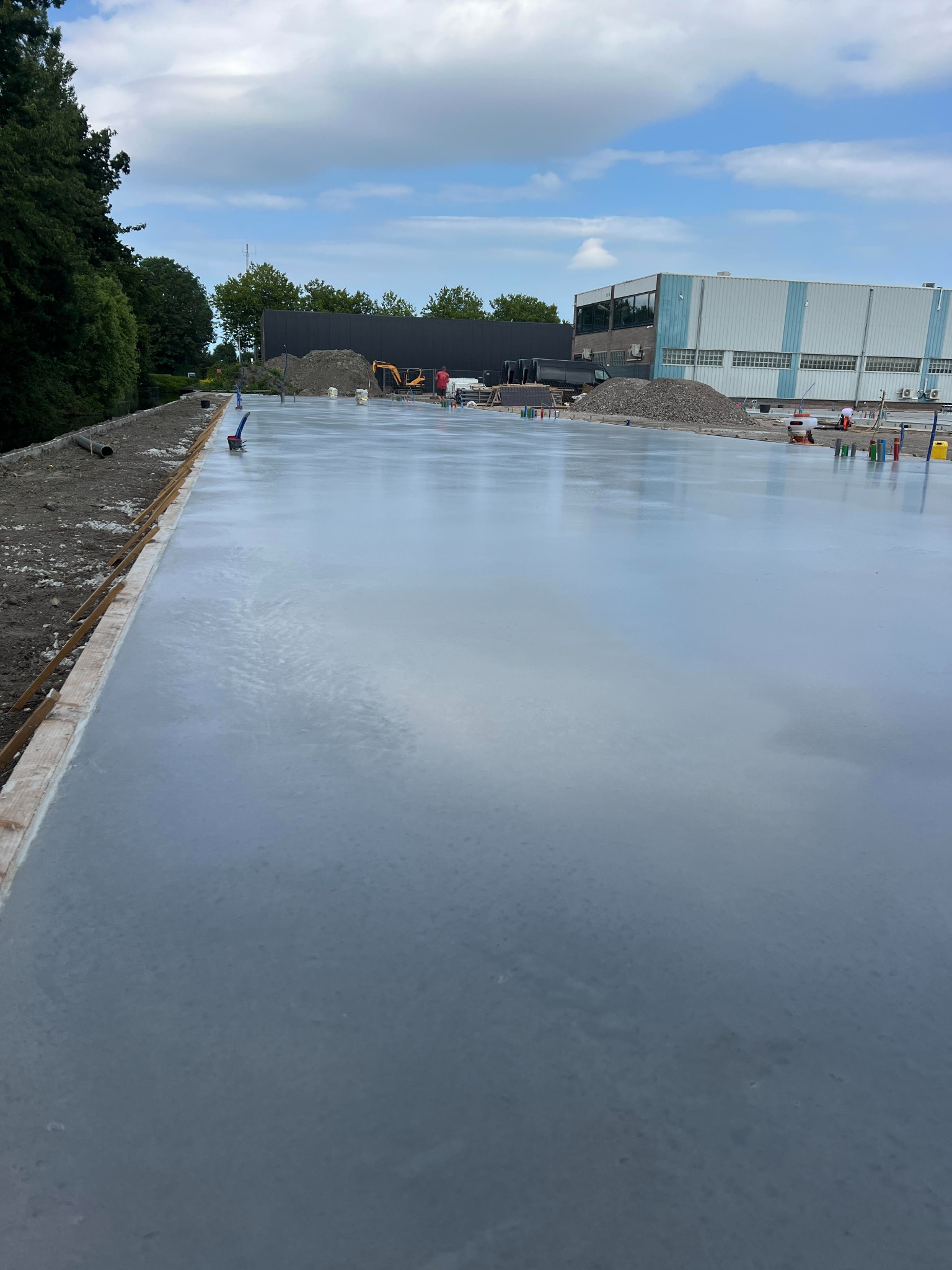 Świeżo wylana, gładka posadzka przemysłowa na zewnątrz budynku, z widocznym sprzętem budowlanym i przygotowanym podłożem. W tle budynek przemysłowy i drzewa.