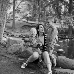 Magdalena Siemaszko Fotografia - Czarno-białe ujęcie matki z dwójką synów siedzących na kamieniach w parku w Gdańsku. Kobieta uśmiecha się, trzyma w ręku aparat fotograficzny. Zdjęcie plenerowe.