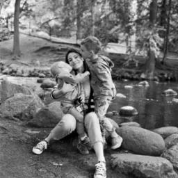 Czarno-białe zdjęcie: Mama siedzi na kamieniach nad rzeką, przytulając dwóch synów. Jeden syn całuje ją w policzek, drugi jest trzymany na kolanach. Aparat na szyi.