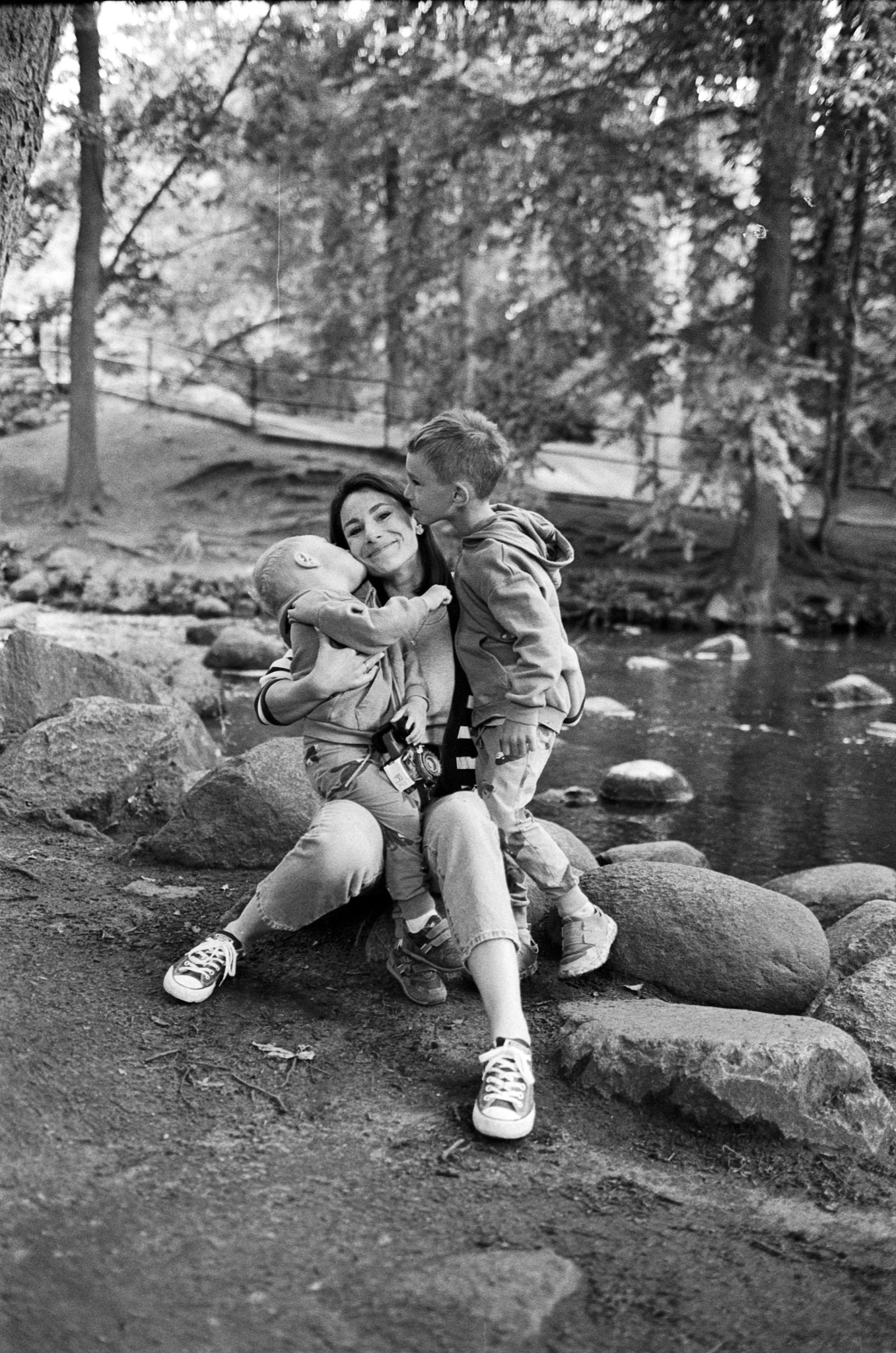 Czarno-białe zdjęcie: Mama siedzi na kamieniach nad rzeką, przytulając dwóch synów. Jeden syn całuje ją w policzek, drugi jest trzymany na kolanach. Aparat na szyi.