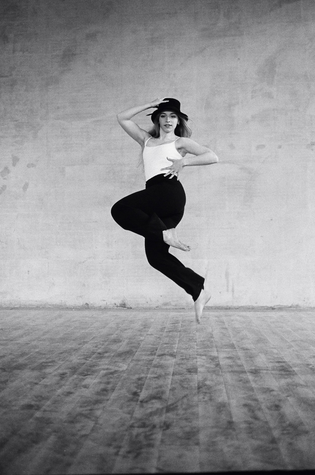 Czarno-białe zdjęcie kobiety w kapeluszu, białym topie i czarnych spodniach, w dynamicznym skoku na tle surowej ściany i drewnianej podłogi. Artystyczna fotografia.