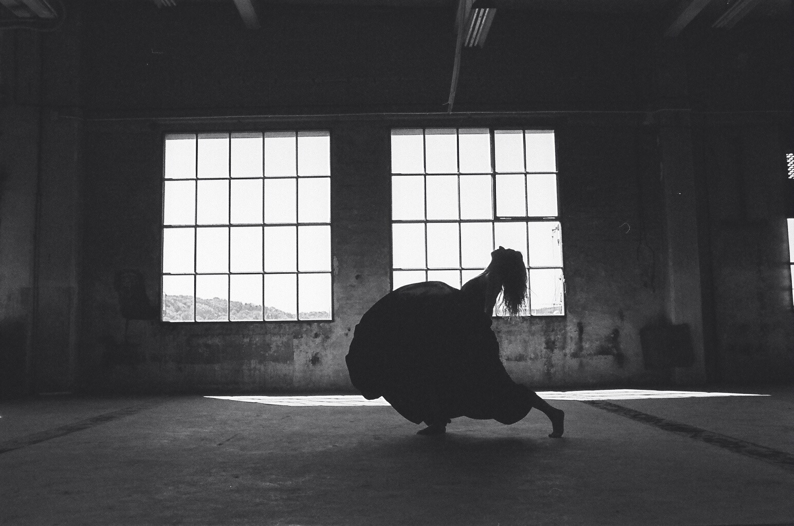 Czarno-białe zdjęcie sylwetki kobiety w tańcu na tle okien w opuszczonym budynku. Mocne kontrasty światła i cienia, surowa estetyka wnętrza, ekspresja ruchu i emocji.